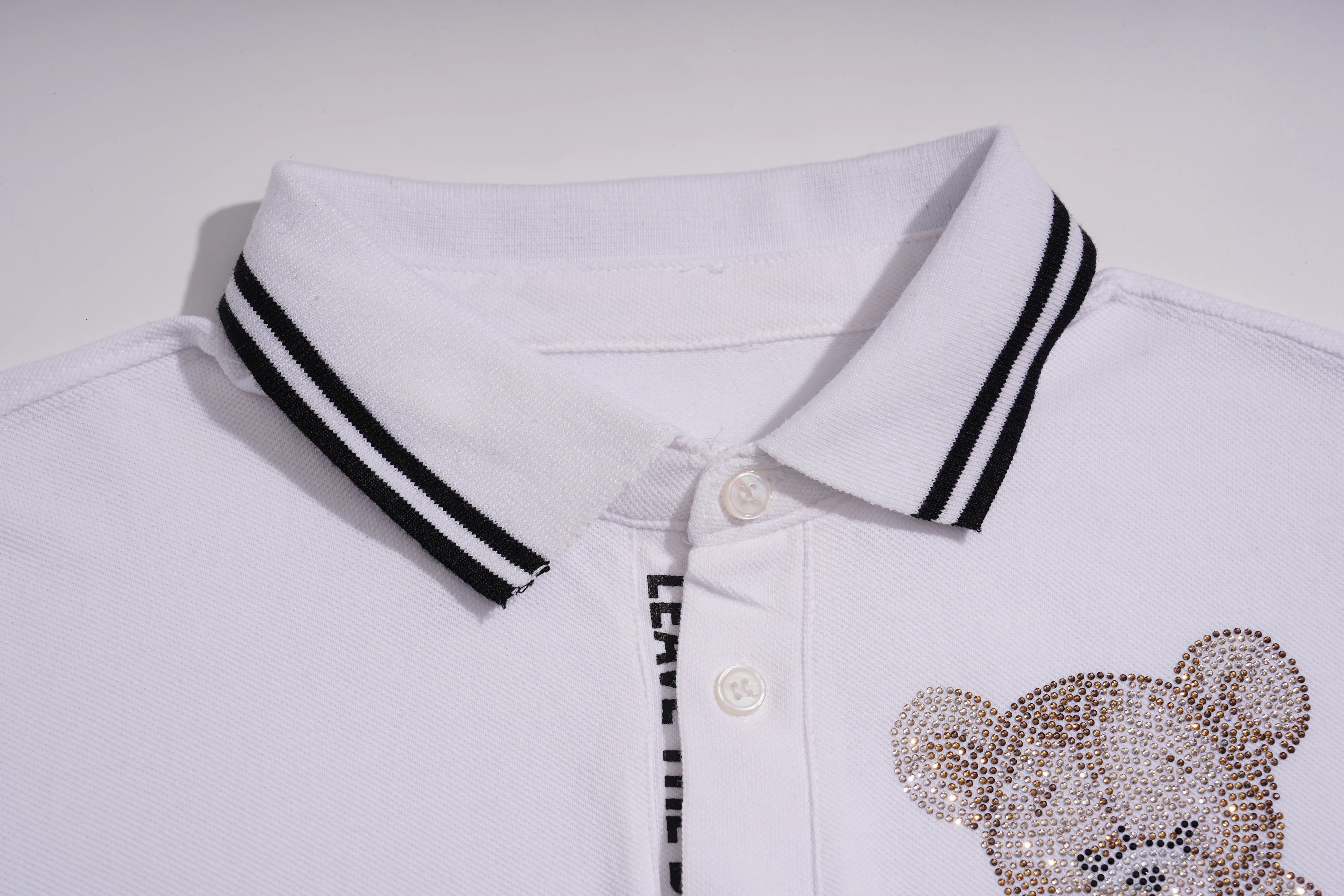 Close-up of a white polo shirt collar with black double stripes, white buttons, and a teddy bear face embellished with rhinestones on the chest. TCH White Teddy Bear Embellished Polo Shirt
