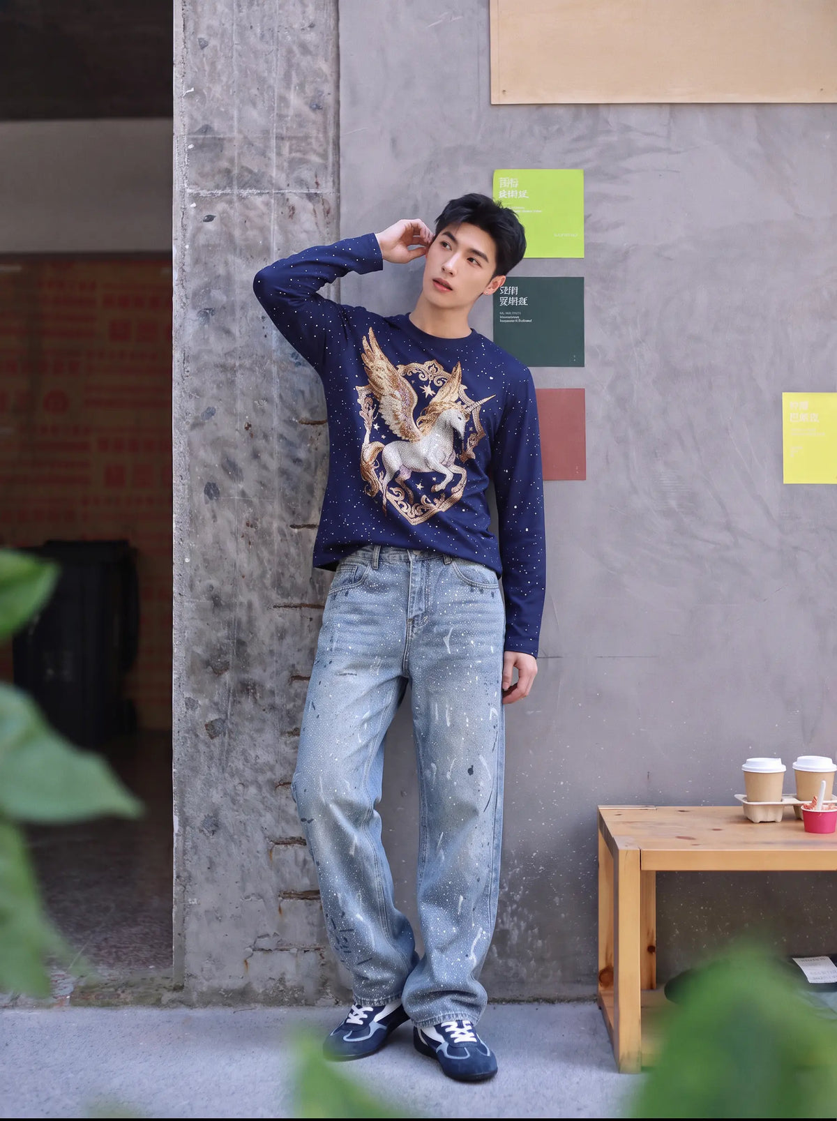 A man wearing a navy blue long-sleeve sweater with a large eagle graphic on the front, paired with light-wash distressed jeans and dark sneakers, posing in front of a textured gray wall. TCH Winged Unicorn Starry Rhinestone Long Sleeve T-Shirt