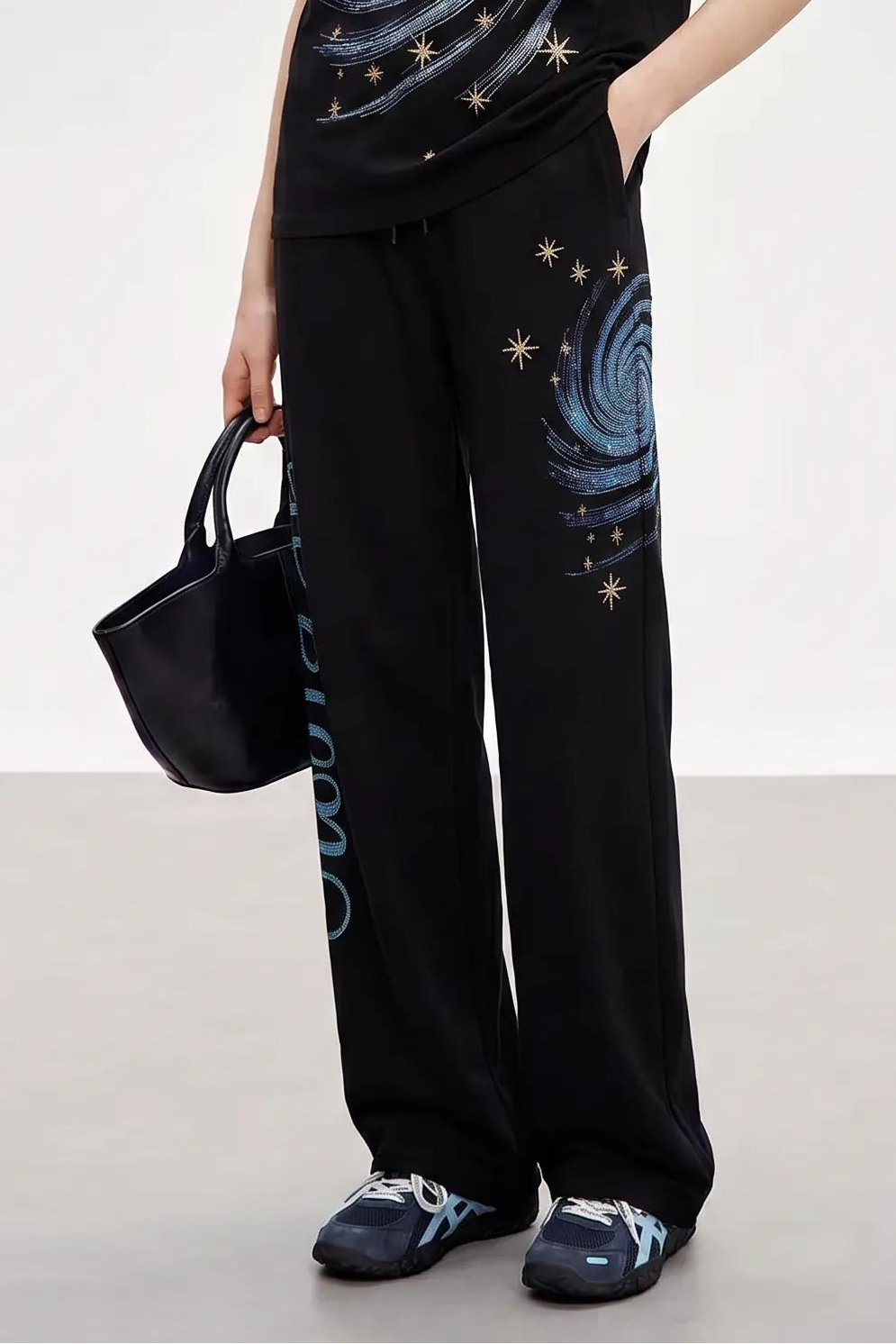 A person wearing black wide-leg sweatpants with a blue swirl and gold star print on the leg, paired with a matching black top and dark sneakers, holding a black handbag against a clean studio background. TCH Women’s Black Wide-Leg Sweatpants with Rhinestone Star Graphic