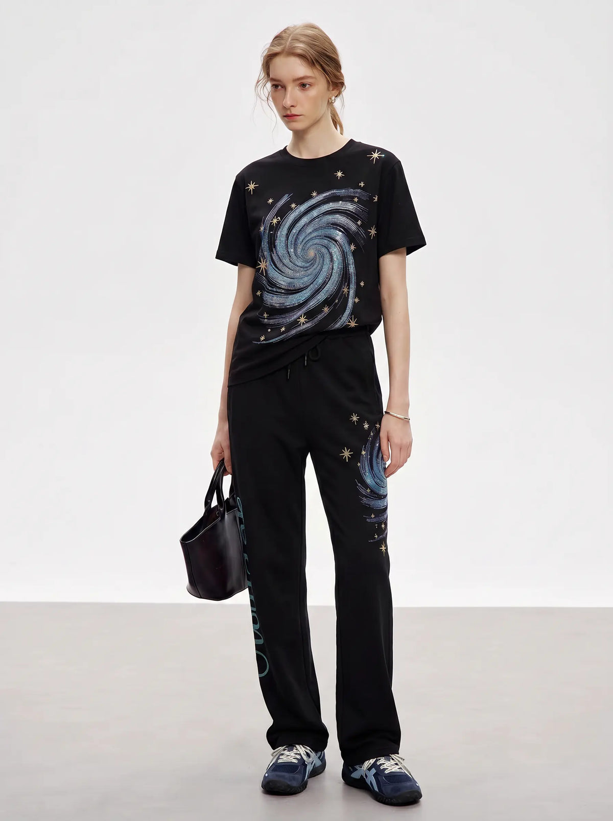 A model stands in a studio wearing a black t-shirt and matching sweatpants with a galaxy swirl print, holding a small black handbag. TCH Women’s Black Wide-Leg Sweatpants with Rhinestone Star Graphic