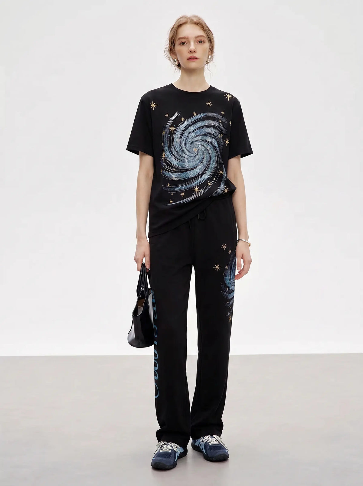 A model stands against a light gray studio background wearing a black short-sleeve T-shirt and matching black pants with galaxy and star print details, along with blue sneakers and carrying a black handbag. TCH Women’s Black Wide-Leg Sweatpants with Rhinestone Star Graphic