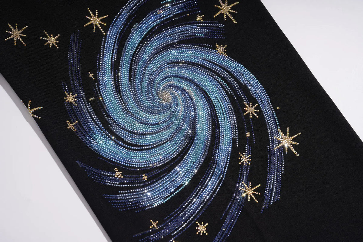 A close-up of a black garment featuring a large spiral galaxy design made of blue and silver rhinestones, accented with small gold star embellishments. TCH Women’s Black Wide-Leg Sweatpants with Rhinestone Star Graphic