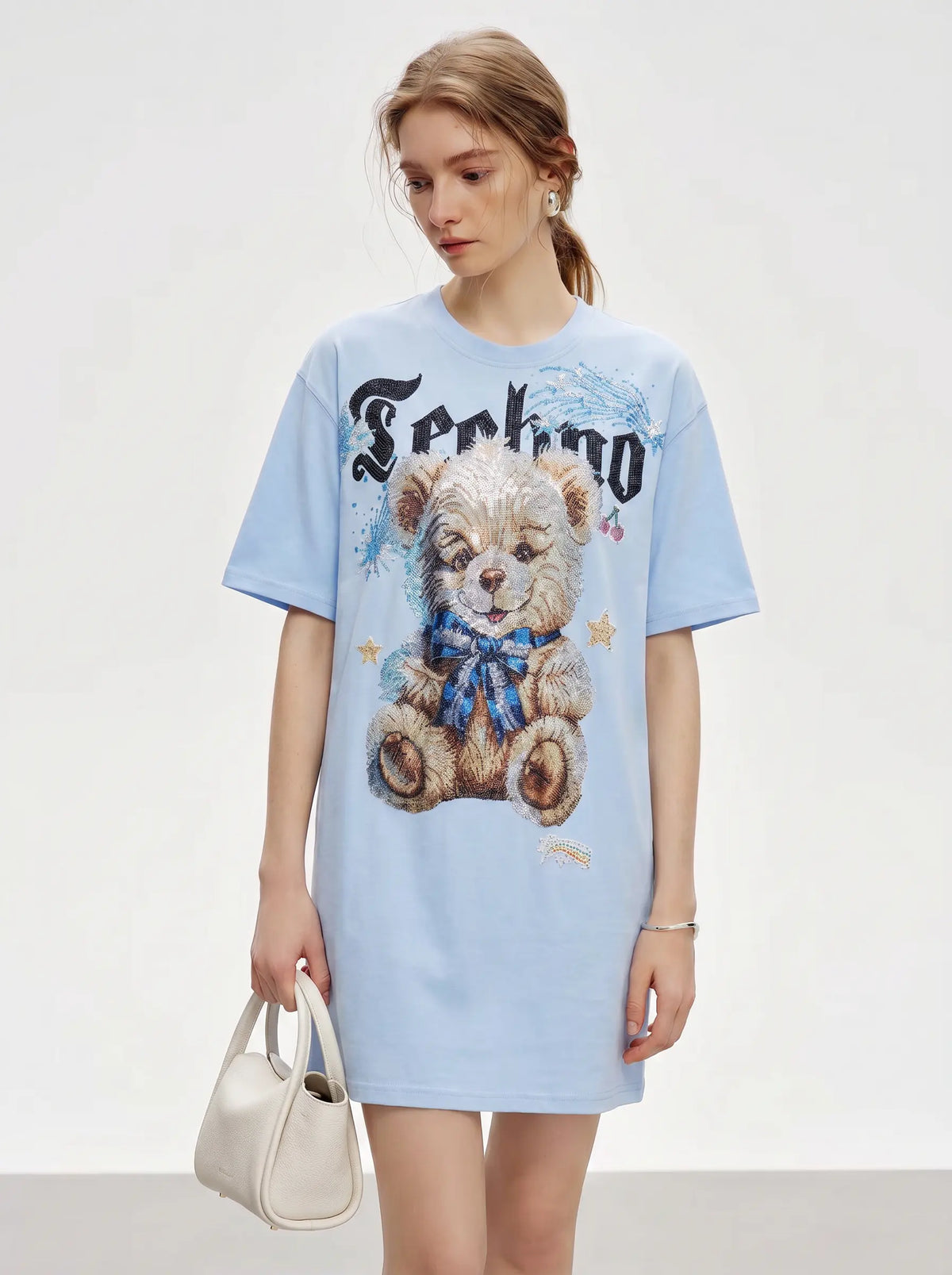 A woman modeling an oversized light blue graphic T-shirt dress featuring a teddy bear illustration and the word 'Sehno' in a gothic font, paired with a small cream handbag against a plain light gray studio background. TCH Women's Blue Teddy Bear Graphic Oversized T-Shirt