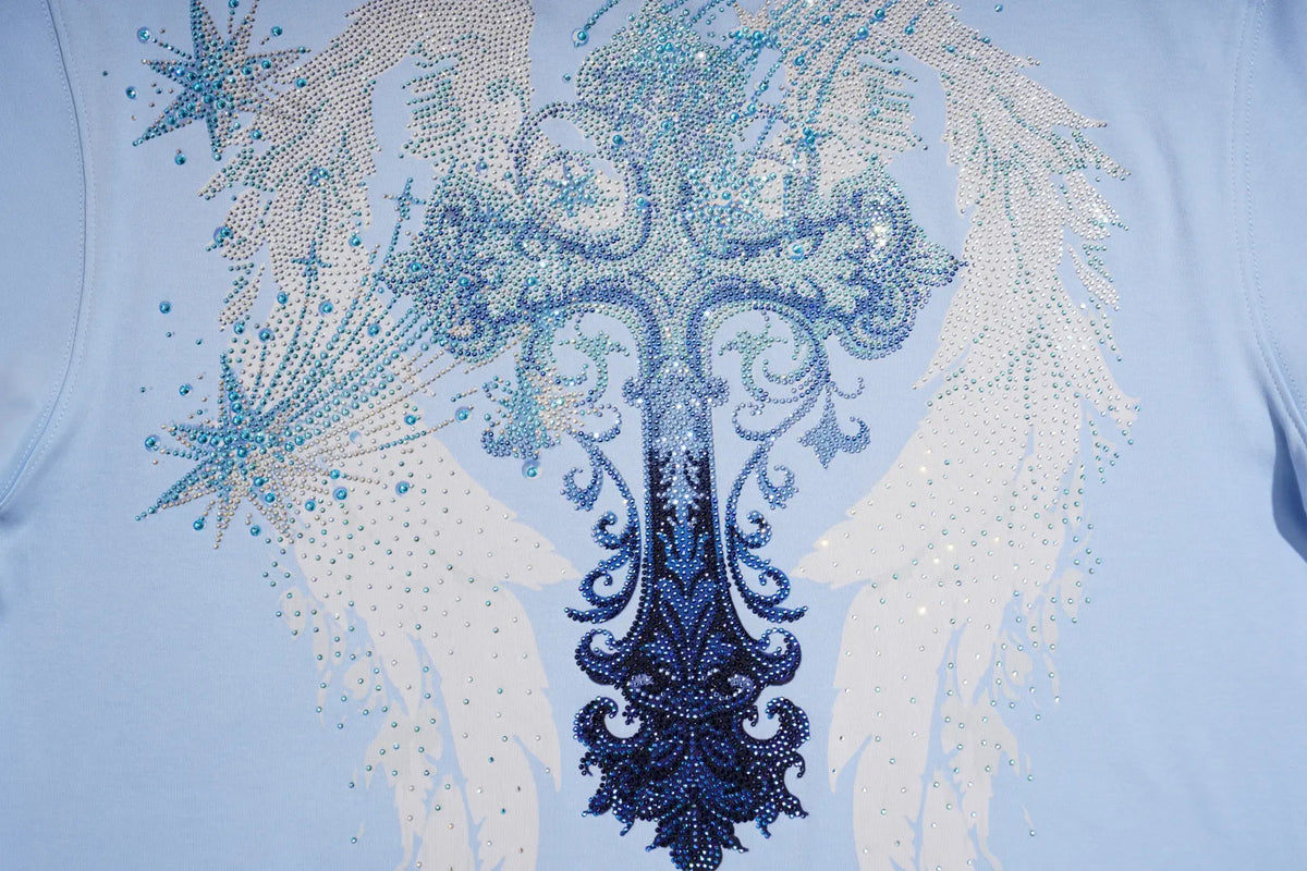 A close-up of a light blue t-shirt featuring a large, ornate graphic design embellished with rhinestones. The design depicts a central dark blue cross-like motif with swirling details, flanked by white angel wings and blue starburst accents. TCH Women's Blue Teddy Bear Graphic Oversized T-Shirt