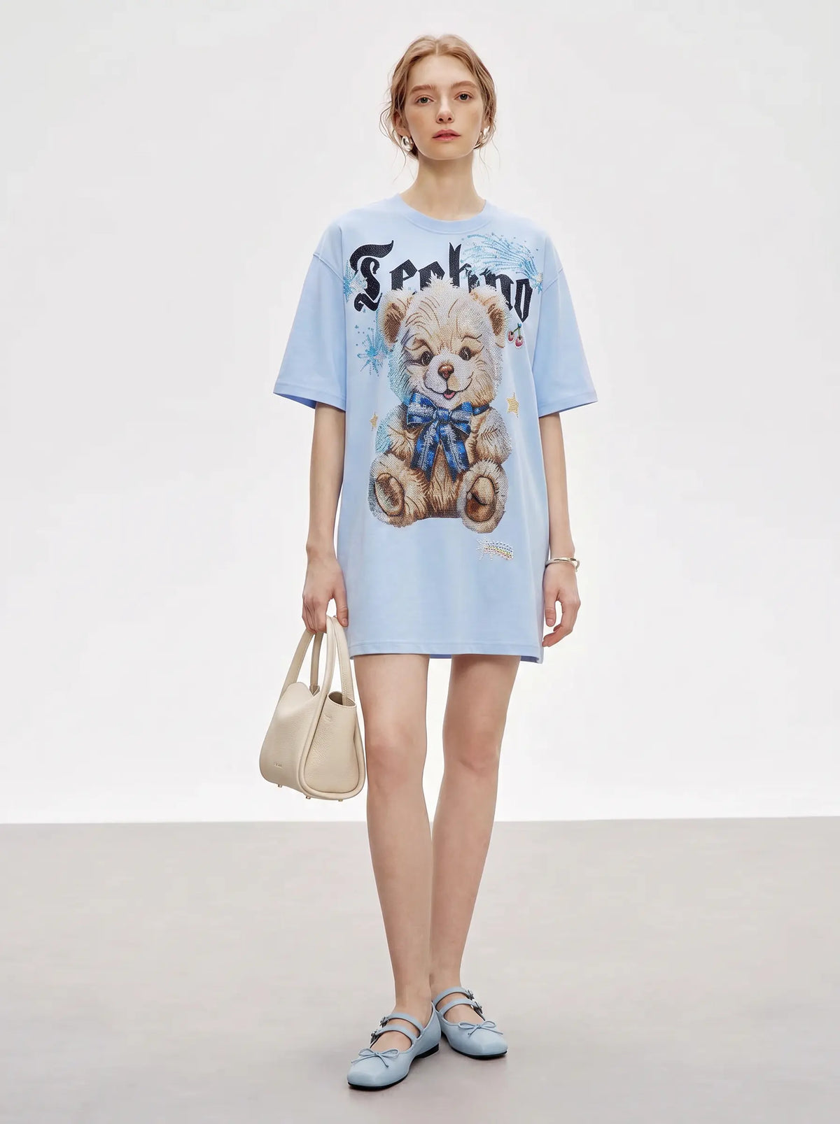 A model stands in a studio wearing a light blue oversized graphic T-shirt dress with a teddy bear print and the word 'Feeling' on the front, paired with a beige handbag and light blue flats. TCH Women's Blue Teddy Bear Graphic Oversized T-Shirt