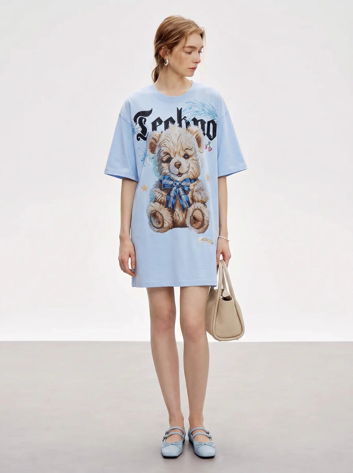 A woman stands in a studio wearing an oversized light blue T-shirt dress with a large teddy bear graphic and the word 'Techno' across the chest, paired with light blue flats and a beige handbag. TCH Women's Blue Teddy Bear Graphic Oversized T-Shirt