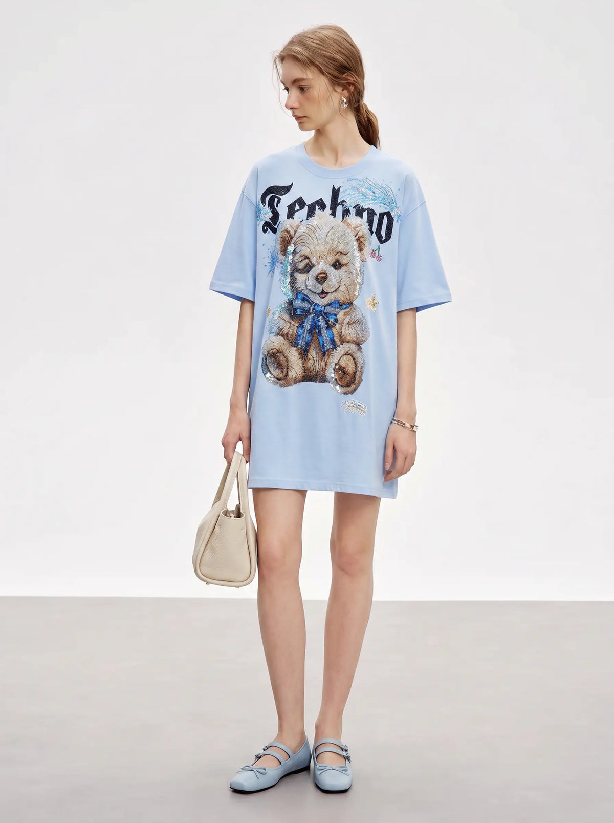 A model stands in a studio wearing an oversized light blue T-shirt dress with a large teddy bear graphic and the word 'Forkno' printed across the chest. She is holding a beige handbag and wearing light blue flats. TCH Women's Blue Teddy Bear Graphic Oversized T-Shirt