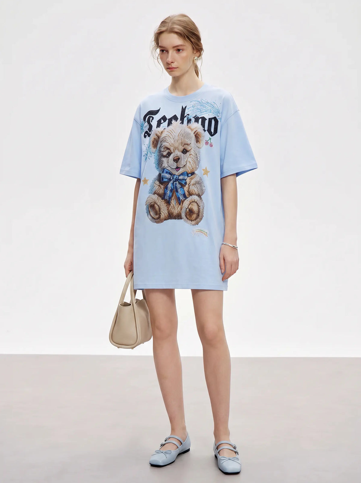 A model stands in a studio wearing an oversized light blue T-shirt dress with a teddy bear graphic and the word 'Ferdmo' across the chest. She is holding a beige handbag and wearing light blue flat shoes against a clean white and gray background. TCH Women's Blue Teddy Bear Graphic Oversized T-Shirt
