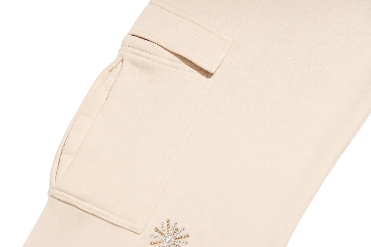 Close-up of a light beige or cream-colored fabric garment, showing a pocket with a flap tab and a small decorative embellishment near the hem. TCH Women’s Cream Cargo Sweatpants with Teddy Bear Rhinestone Graphic