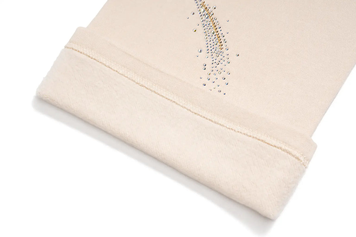 A close-up of a cream-colored knit garment, likely a sweater or scarf, featuring a folded cuff with a neat hem and a decorative vertical line of small metallic rhinestones in gold and silver. TCH Women’s Cream Cargo Sweatpants with Teddy Bear Rhinestone Graphic
