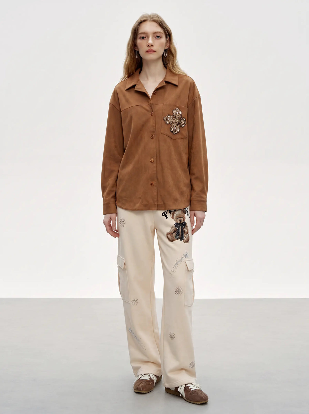 A model stands against a light gray studio background wearing a brown long-sleeve button-up shirt with a floral applique on the chest pocket, cream cargo pants with subtle floral prints, and brown slip-on shoes. TCH Women’s Cream Cargo Sweatpants with Teddy Bear Rhinestone Graphic