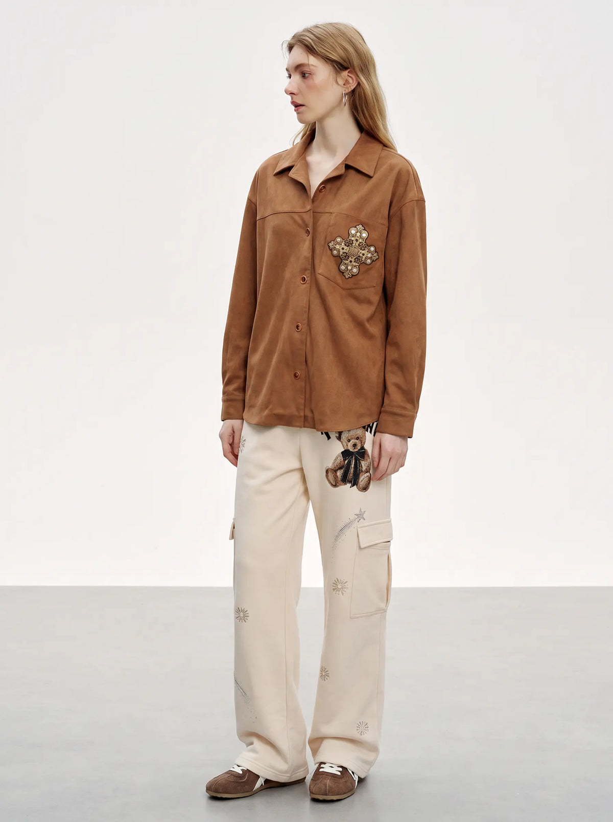 A model stands in a studio wearing a brown button-up shirt with a floral embroidered patch on the pocket, paired with cream cargo pants and brown shoes. TCH Women’s Cream Cargo Sweatpants with Teddy Bear Rhinestone Graphic