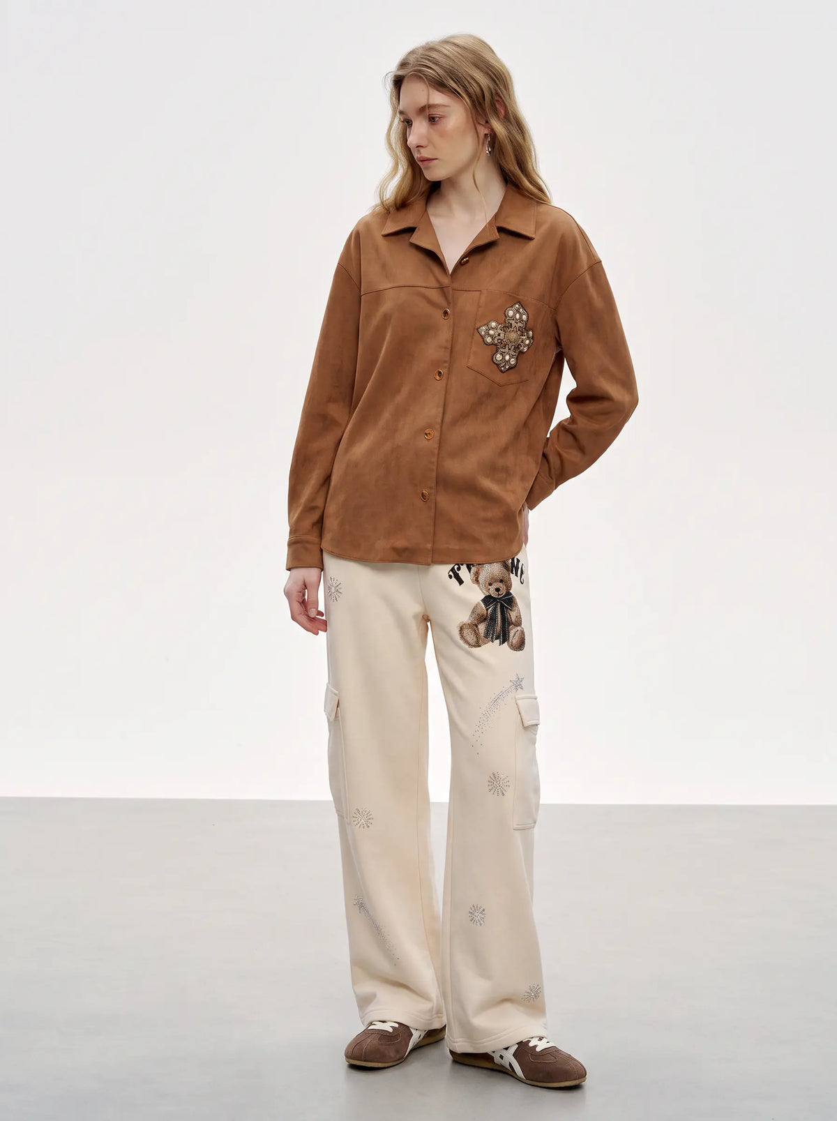 A model stands in a studio wearing a brown suede-like button-up shirt with a floral applique on the chest pocket, paired with cream cargo pants featuring teddy bear embroidery and brown slip-on shoes. TCH Women’s Cream Cargo Sweatpants with Teddy Bear Rhinestone Graphic