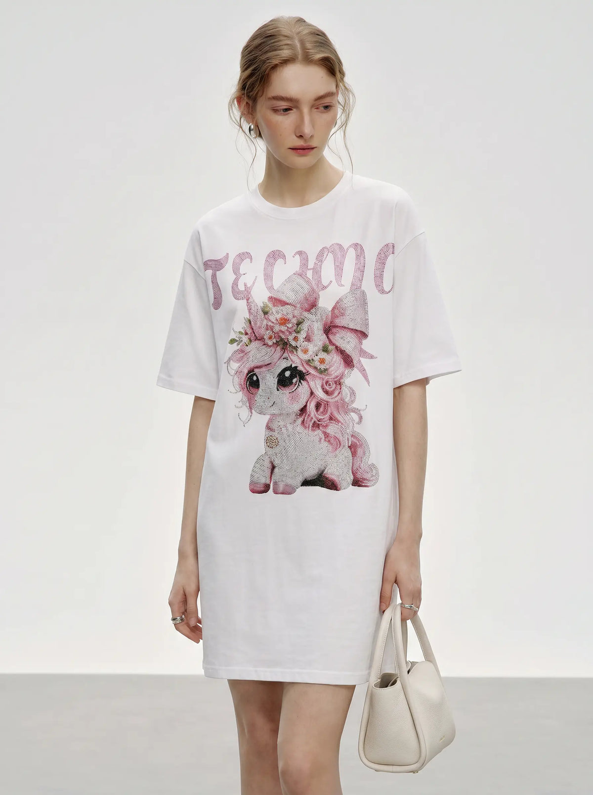A model wearing a white oversized T-shirt dress with a pink sequined graphic of a cartoon pony and the word 'TEACH' in pink sequins, holding a small cream-colored handbag against a light gray studio background. TCH Women's Pink Angel Graphic Oversized T-Shirt