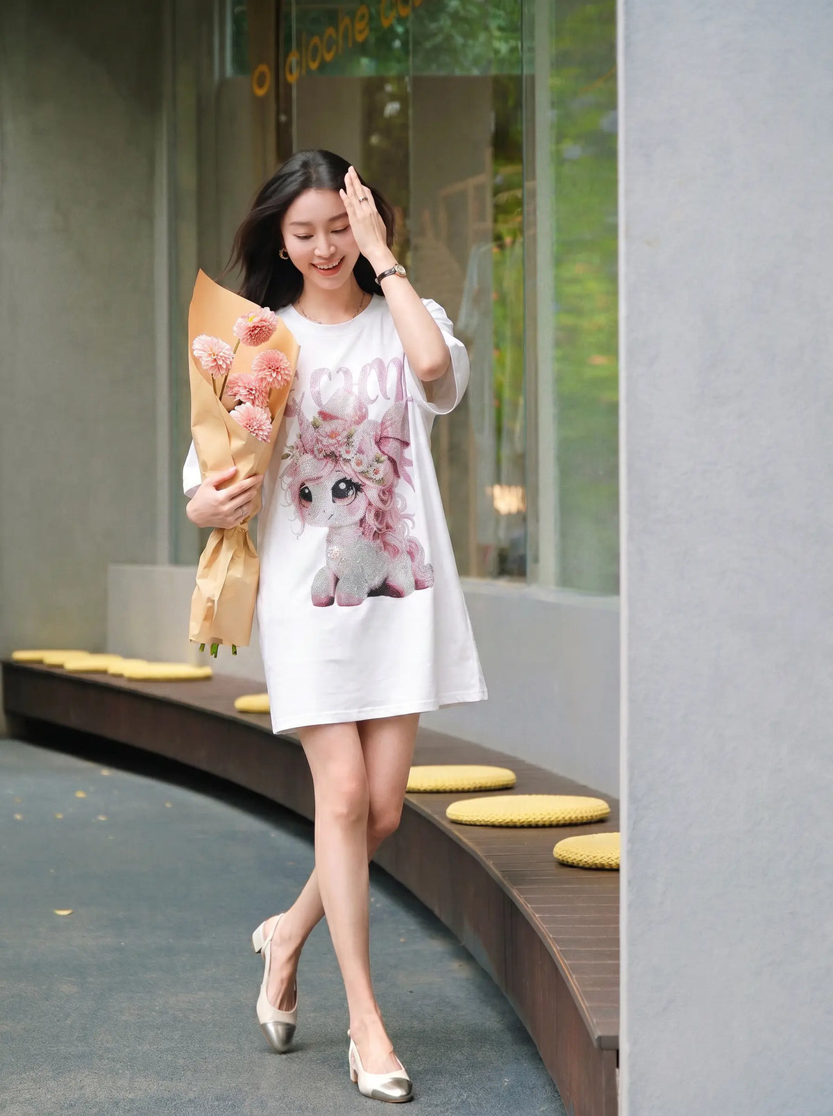 A woman walking outdoors while wearing a white oversized graphic T-shirt dress with a pink cartoon cat print and holding a bouquet of pink flowers. TCH Women's Pink Angel Graphic Oversized T-Shirt