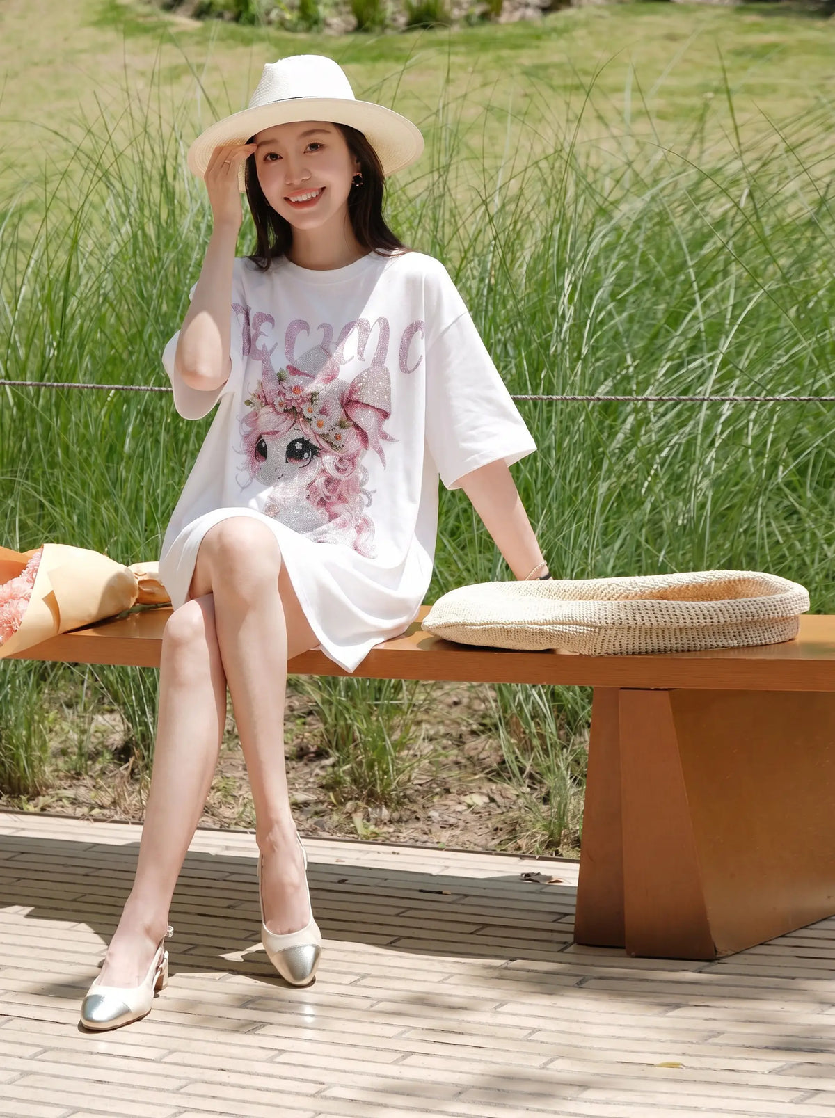 A woman sits on a wooden bench outdoors wearing a white oversized graphic T-shirt dress with a pink floral print, a white wide-brim hat, and metallic gold shoes. Tall green grass and a paved walkway are visible in the background. TCH Women's Pink Angel Graphic Oversized T-Shirt