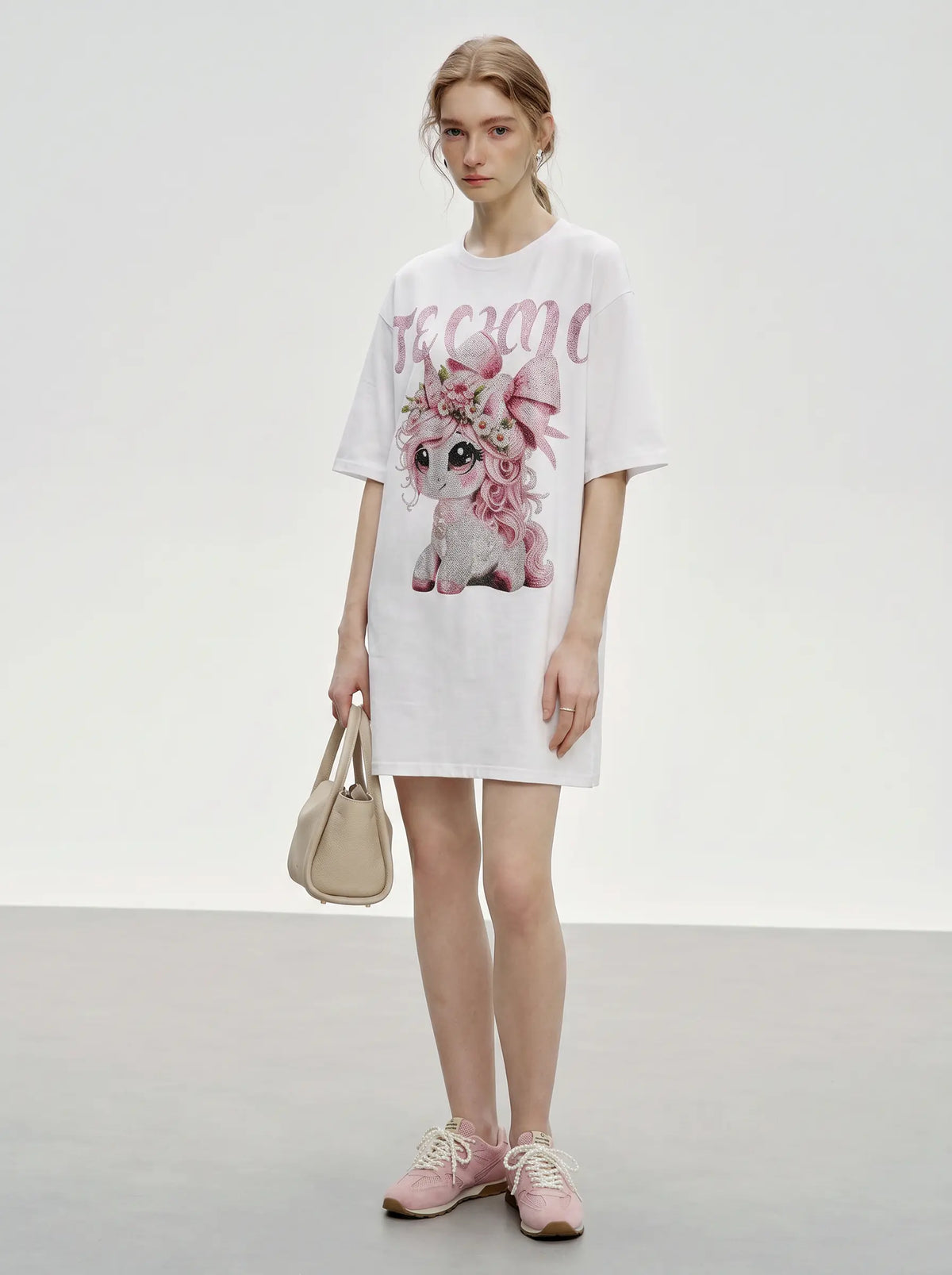 A model stands in a studio wearing an oversized white T-shirt dress with a pink graphic of a cartoon dog and the word 'dream' in pink script. She is holding a beige handbag and wearing pink sneakers. TCH Women's Pink Angel Graphic Oversized T-Shirt