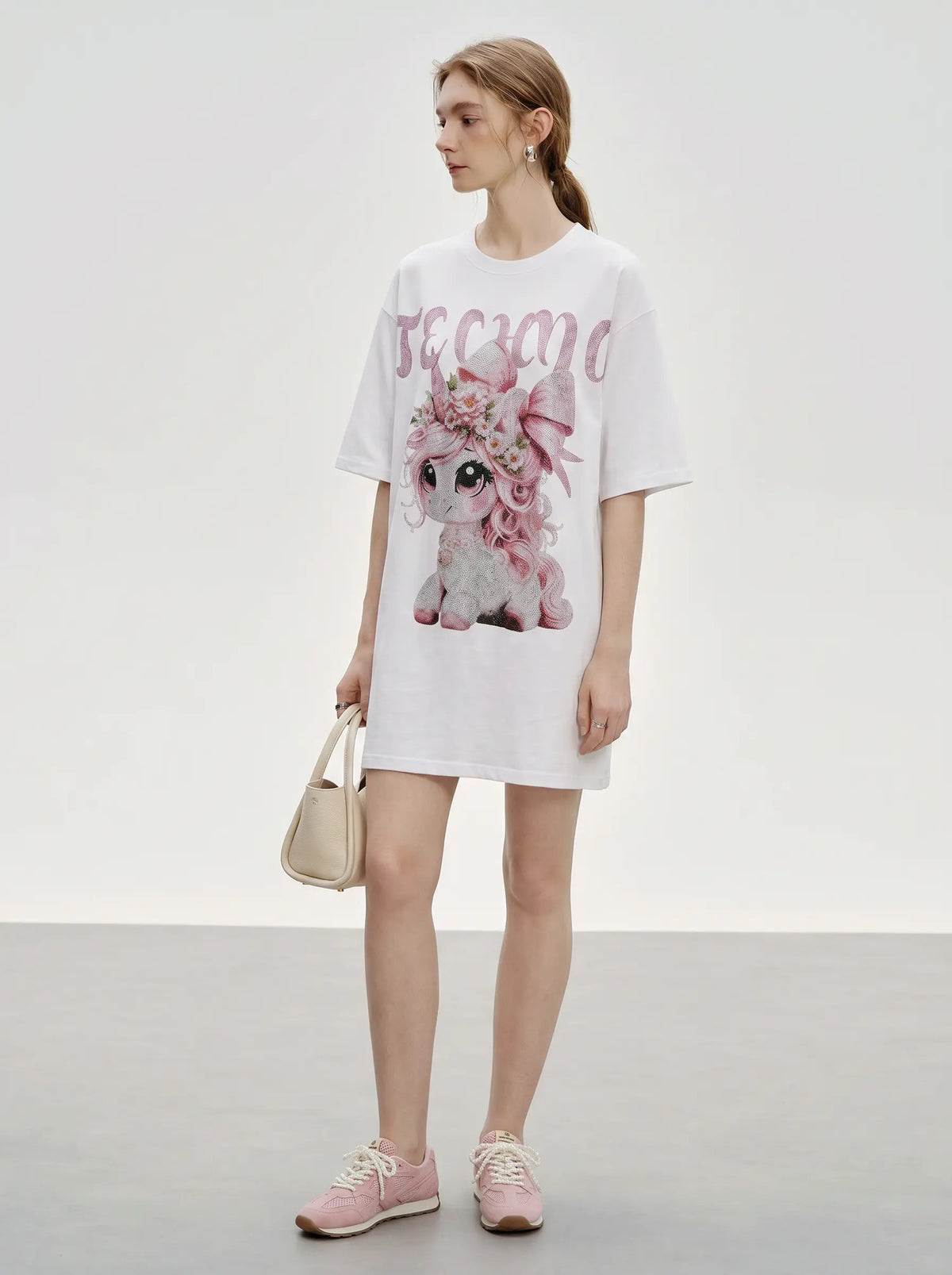 A woman modeling a white oversized graphic T-shirt dress with a pink cartoon dog and the word 'BEACH' in pink letters, paired with pink sneakers and a small beige handbag against a plain studio background. TCH Women's Pink Angel Graphic Oversized T-Shirt