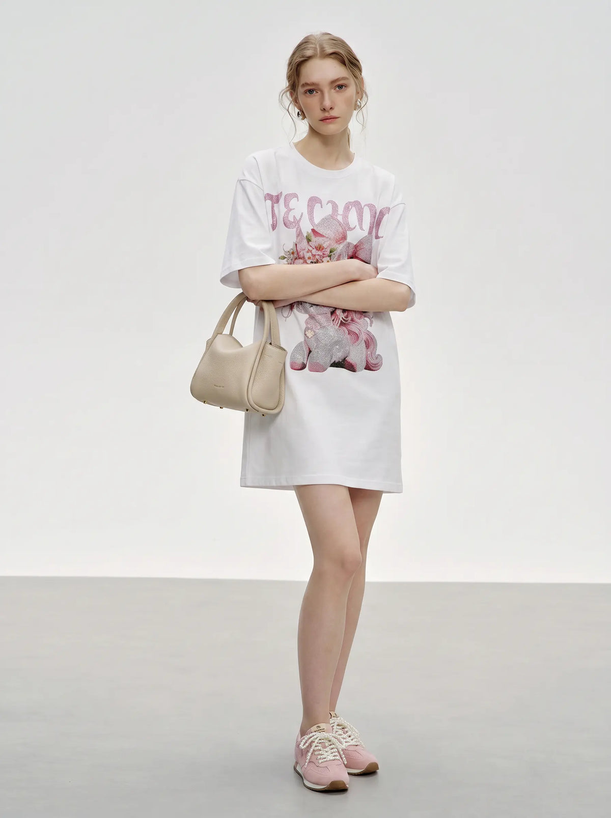 A model stands in a studio wearing a white oversized T-shirt dress with a pink graphic print, holding a beige handbag and wearing pink sneakers. TCH Women's Pink Angel Graphic Oversized T-Shirt