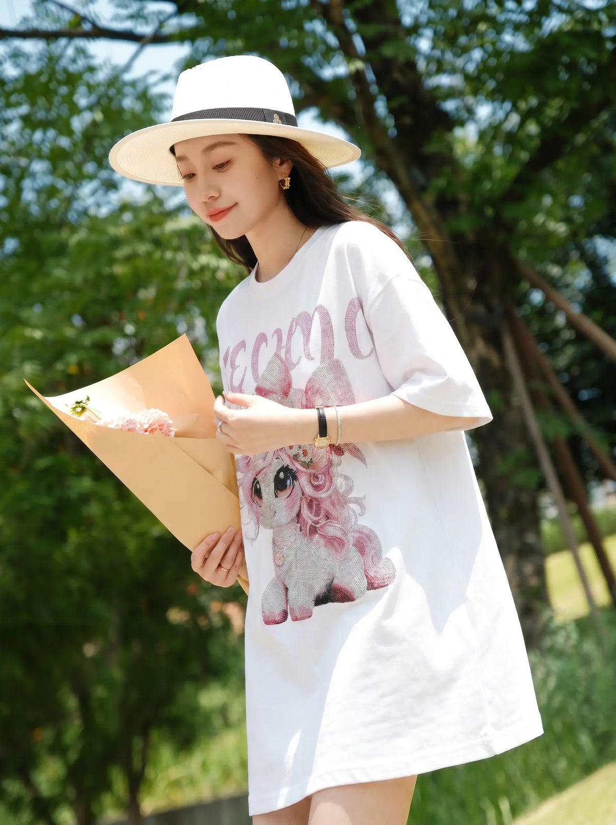 A woman wearing a white oversized graphic T-shirt dress with a pink unicorn print and 'EAMO' lettering, paired with a white fedora hat and holding a bouquet of flowers in an outdoor park setting. TCH Women's Pink Angel Graphic Oversized T-Shirt