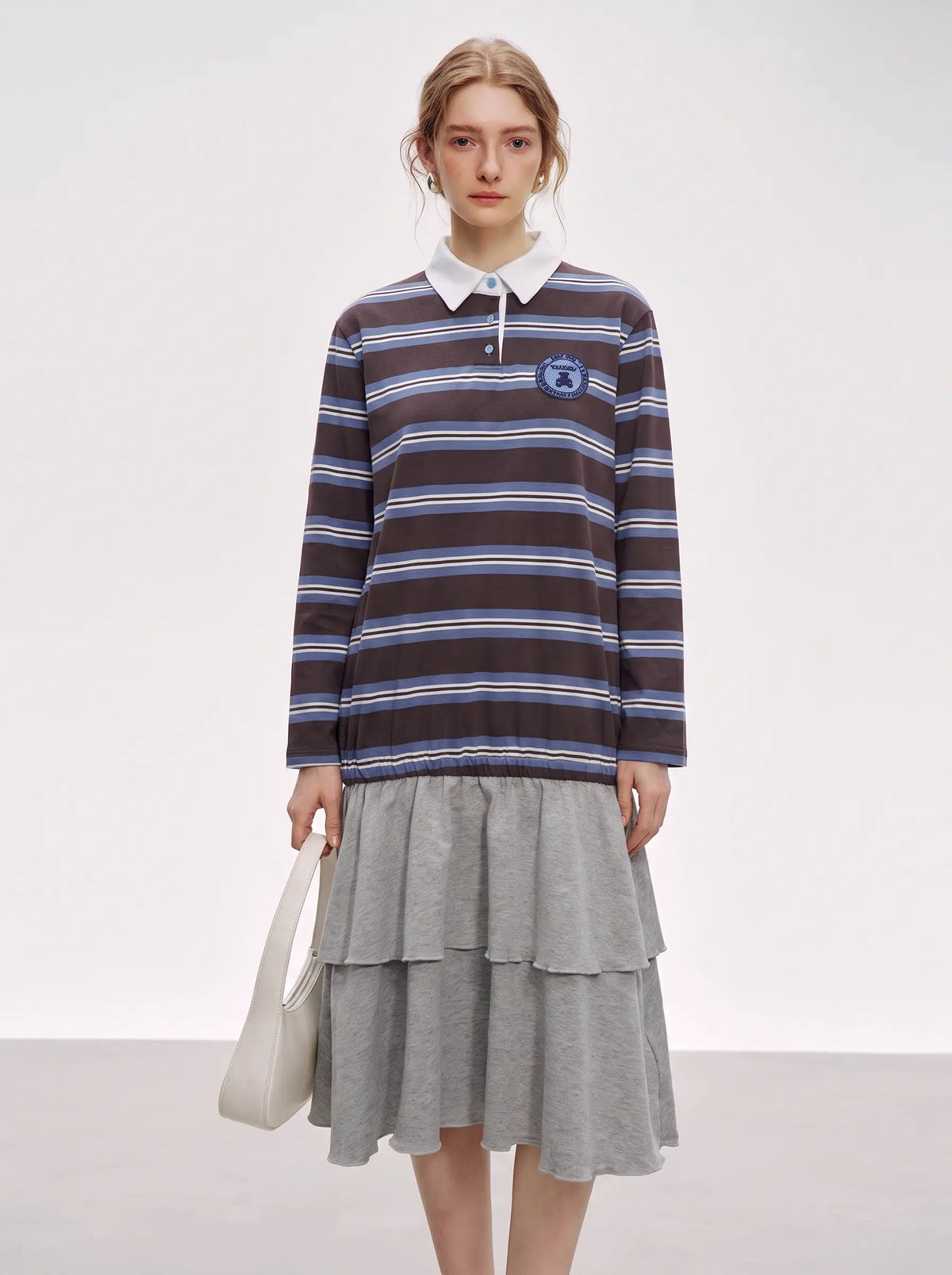 A model stands against a light gray studio background wearing a long-sleeve polo-style knit top with horizontal navy, brown, and white stripes, layered over a light gray tiered skirt. She holds a white shoulder bag. TCH Women's Striped Polo Mini Dress