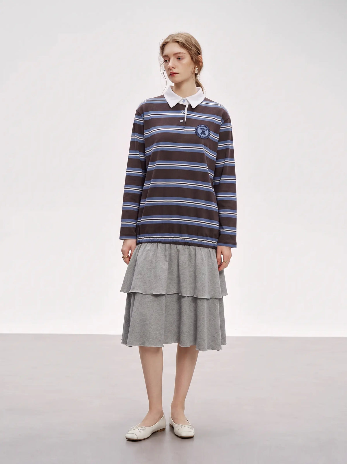 A model stands in a studio wearing a dark brown and blue striped long-sleeve polo shirt with a white collar, paired with a gray tiered skirt and white shoes. TCH Women's Striped Polo Mini Dress