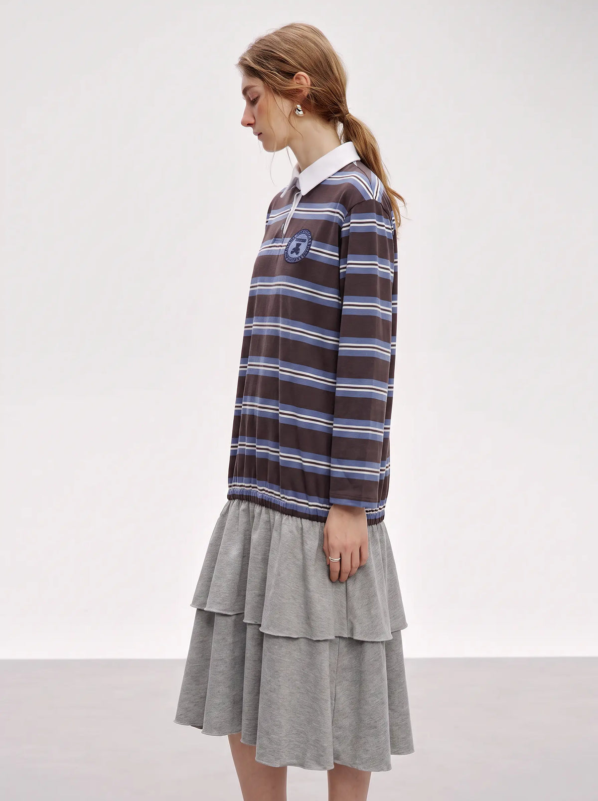 A woman modeling a brown and blue striped long-sleeve rugby shirt with a white collar, paired with a gray tiered skirt, shown from a side profile against a plain light gray studio background. TCH Women's Striped Polo Mini Dress
