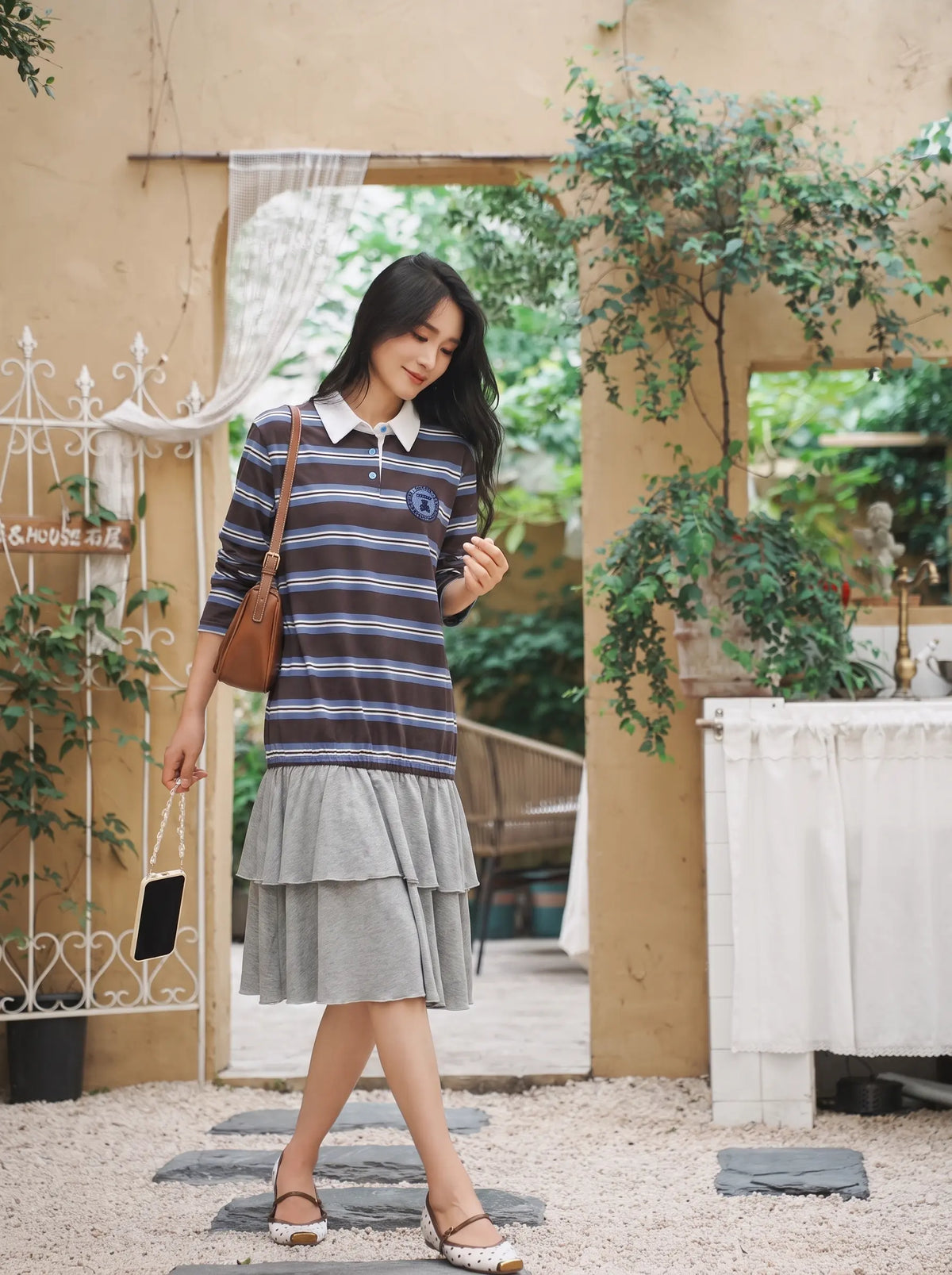 A woman wearing a dark blue and gray striped long-sleeve top with a white collar and a layered light gray skirt, walking in a garden courtyard setting. TCH Women's Striped Polo Mini Dress