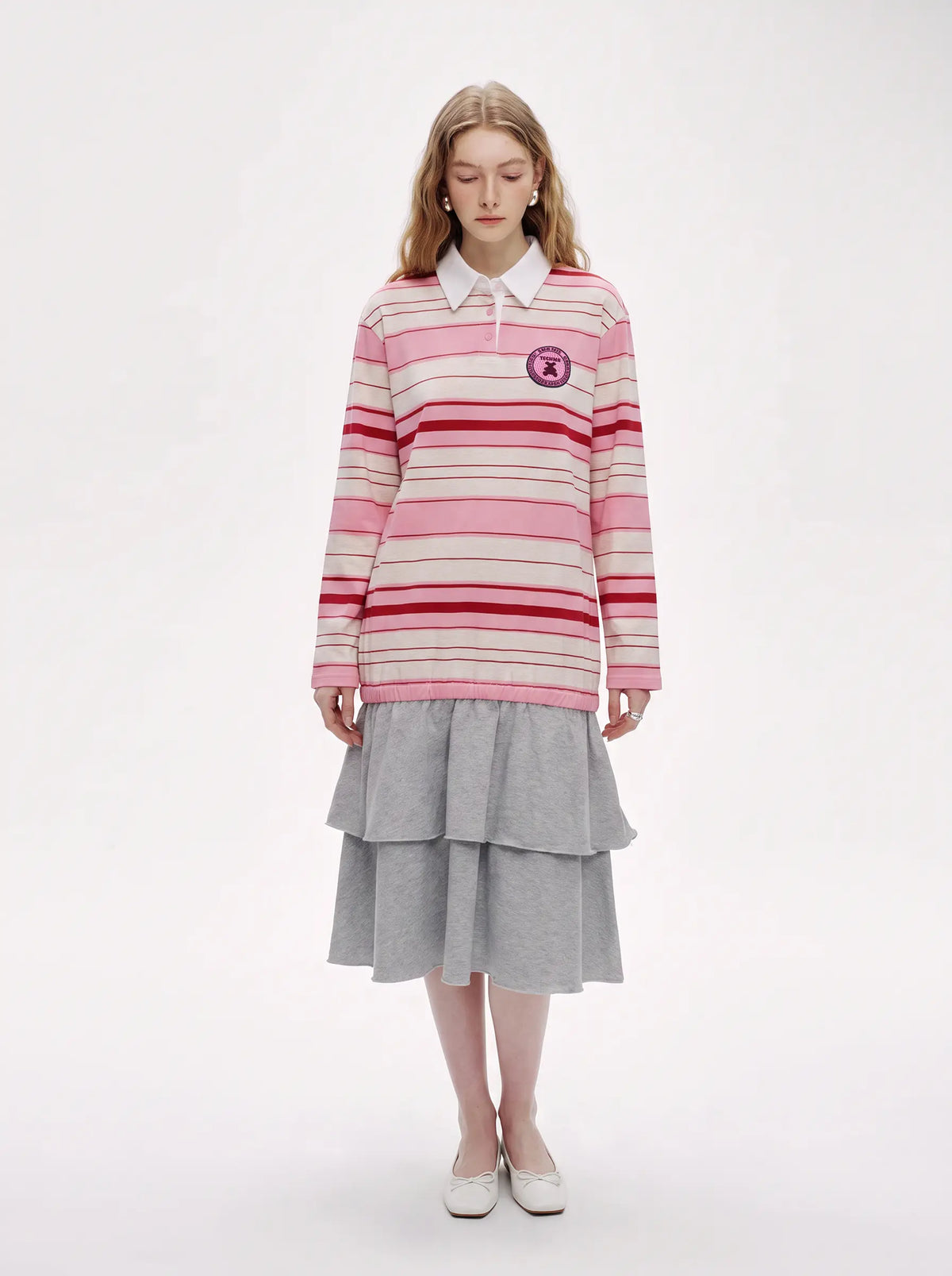 A model stands against a plain white studio background wearing a pink and red striped long-sleeve polo sweater with a white collar, paired with a gray tiered skirt and white shoes. TCH Women's Striped Polo Mini Dress