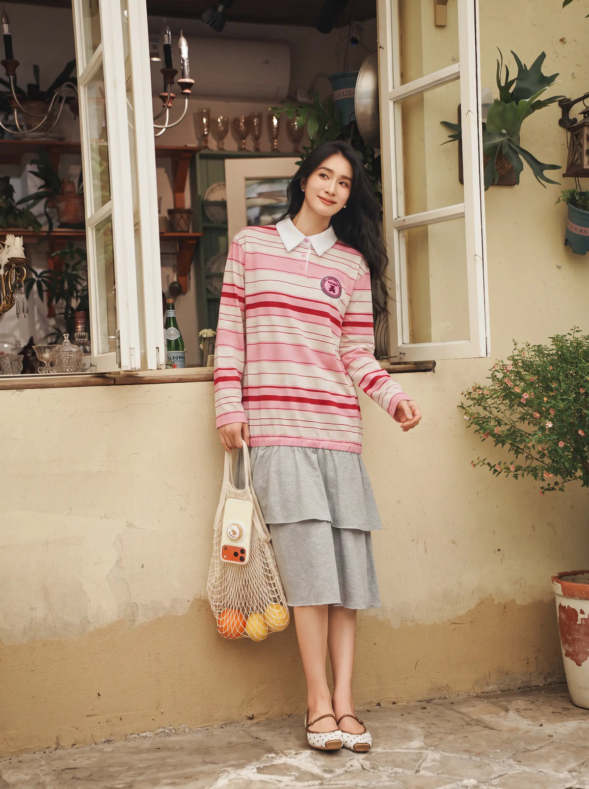 A woman stands outdoors in front of a rustic beige wall and window, wearing a pink and red striped long-sleeve polo shirt with a gray tiered midi skirt. She is holding a beige mesh bag containing fruit and a small bottle. TCH Women's Striped Polo Mini Dress