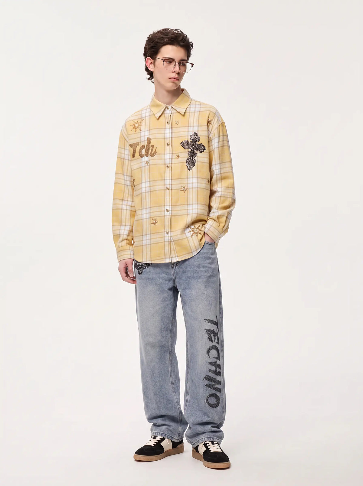 A model stands against a plain white studio background wearing a yellow and white plaid button-up shirt with graphic patches, light-wash blue jeans with 'TECHNO' printed down the leg, and black sneakers. TCH Yellow Plaid Embroidered Cross Shirt