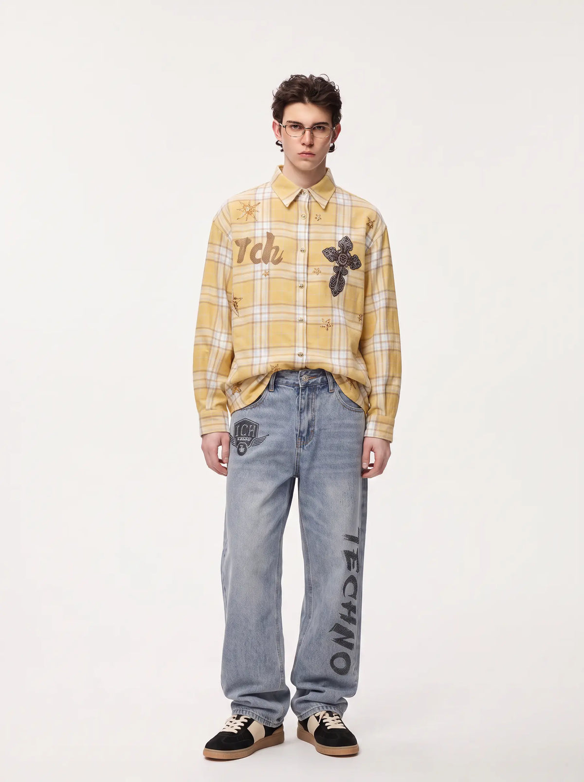 A male model stands against a plain light background wearing a yellow and white plaid button-up shirt with graphic patches, light-wash blue jeans with vertical black lettering, and black sneakers. TCH Yellow Plaid Embroidered Cross Shirt