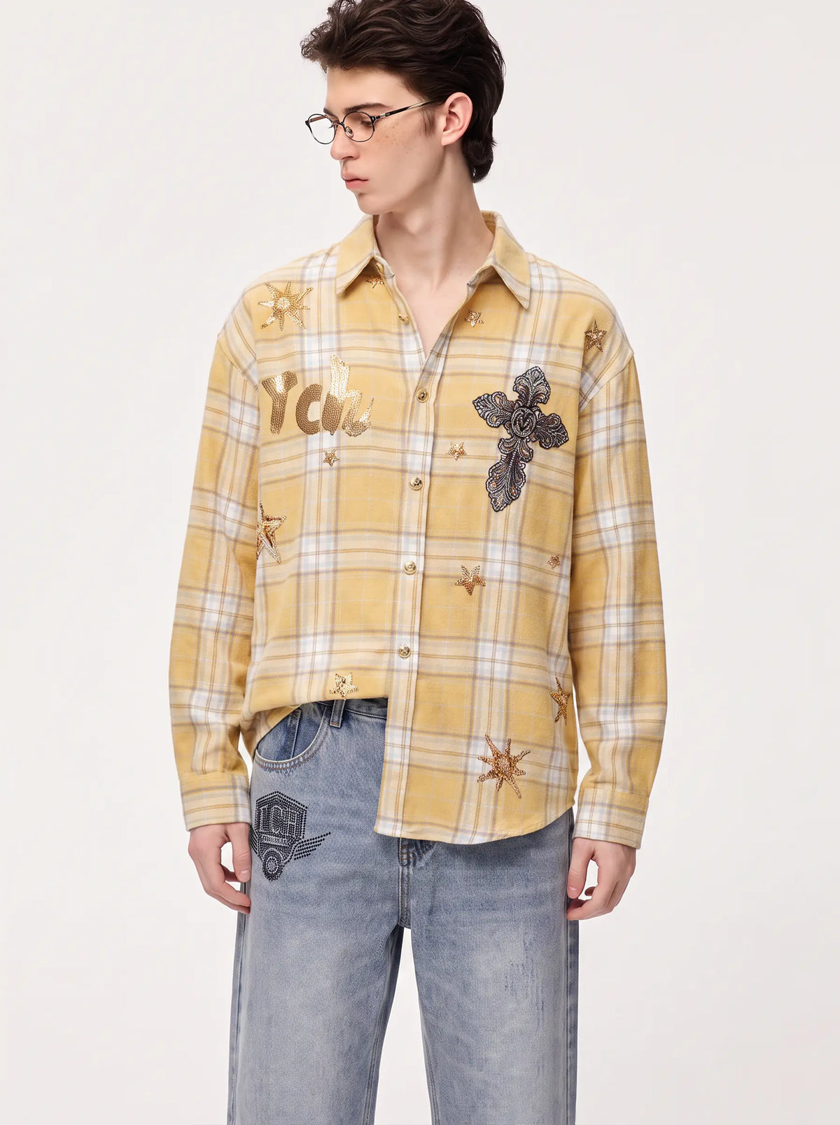 A young man wearing glasses, a light blue and white checkered long-sleeve button-down shirt, a blue patterned tie, and light-wash jeans, standing against a plain light gray studio background. TCH Yellow Plaid Embroidered Cross Shirt