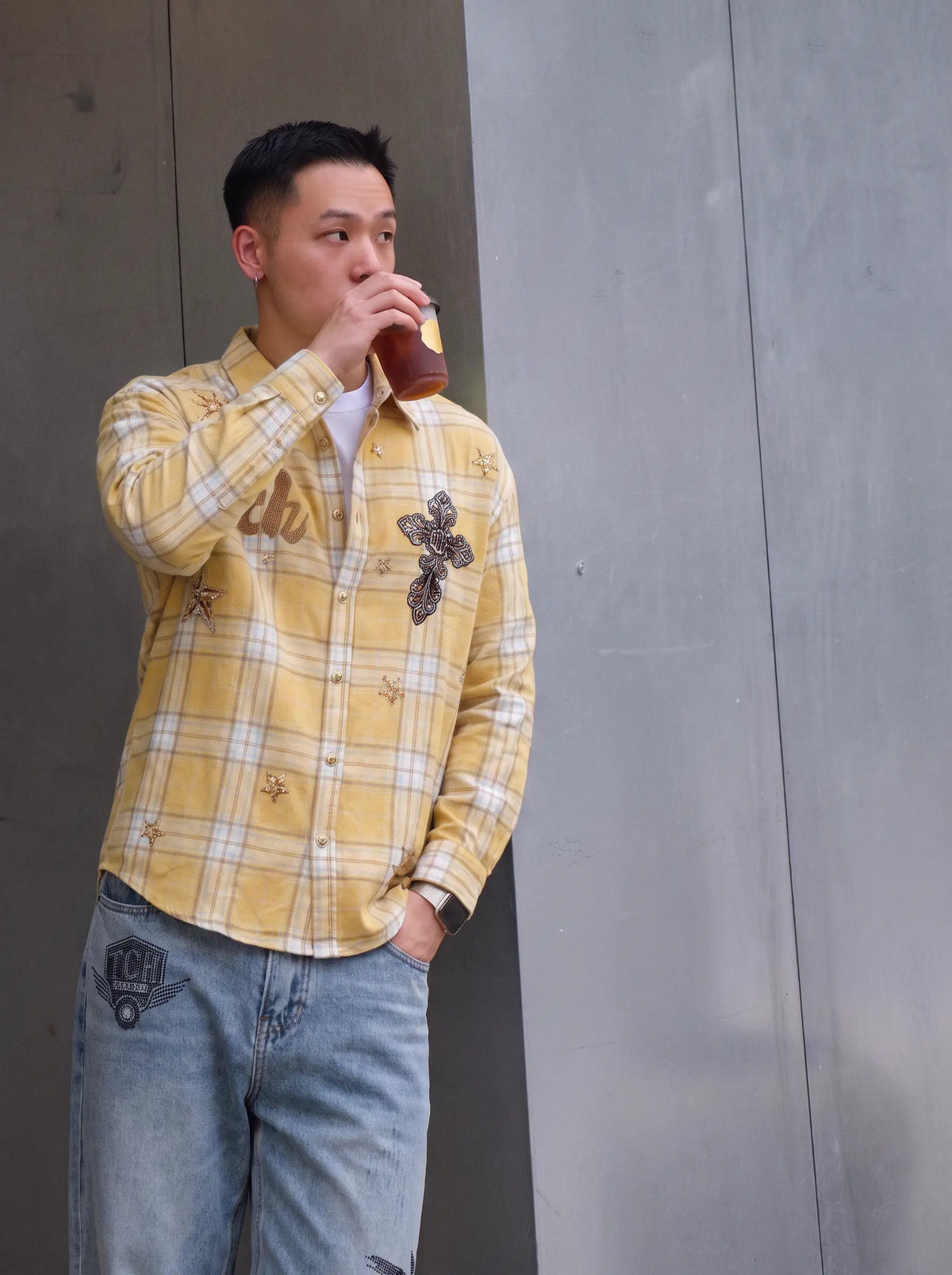 A man wearing a yellow and white plaid button-up shirt with butterfly and bee embroidery, paired with light blue jeans, standing against a gray concrete wall and drinking from a brown bottle. TCH Yellow Plaid Embroidered Cross Shirt